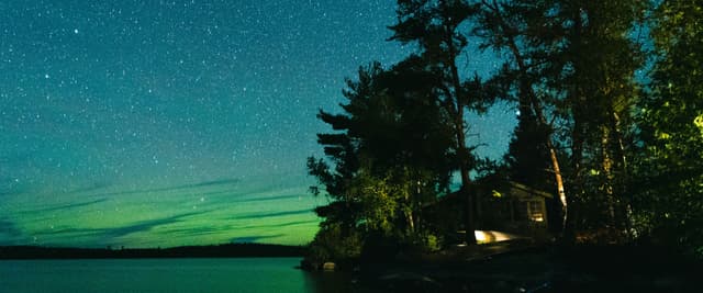 Lakeside cabin at night under a starry sky with northern lights over the water